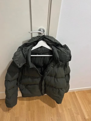 Mörkgrön pufferjacka från Zara - Säljer en mörkgrön pufferjacka från Zara med huva och knappar framtill. Jackan har en schysst look och är perfekt för kalla dagar. Materialet är syntet och jackan har långa ärmar. Grym att ha när det är kallt ute!