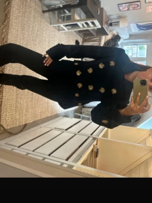 Svart skepparkavaj med guldknappar - Säljer en svart skepparkappa med stora knappar framtill och på ärmsluten. Jackan har en klassisk dubbelknäppt design och är långärmad med bred krage. Materialet ser ut att vara ull, vilket gör den både snygg och varm. Perfekt för dig som gillar en stilren look. Köpt i london