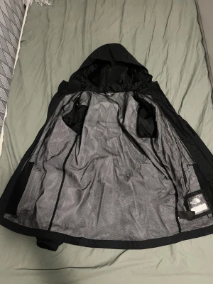 Svart regnjacka från The North Face - Svart regnjacka från The North Face med huva och DryVent-teknologi. Jackan har lång ärm, dragkedja framtill och meshfoder på insidan. Perfekt för blöta dagar och har en clean look med logga på bröstet. Passar XS men också folk som har S beror på lite