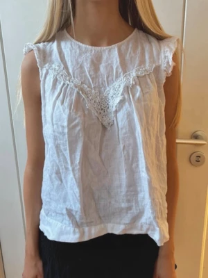 Vit blus med spetsdetaljer från Zara - Säljer en söt vit blus från Zara med spetsdetaljer framtill och volang vid ärmslut. Blusen är ärmlös och har en liten knapp i nacken.