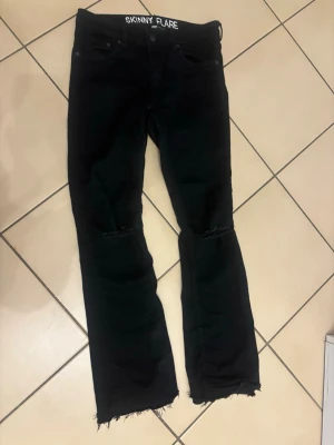 Svarta skinny flare jeans - Svarta jeans med skinny flare-passform och råa, fransiga benslut. Jeansen har slitna detaljer vid knäna och klassisk femficksdesign. Materialet är jeans och färgen är helsvart, vilket ger en cool och edgy vibe.