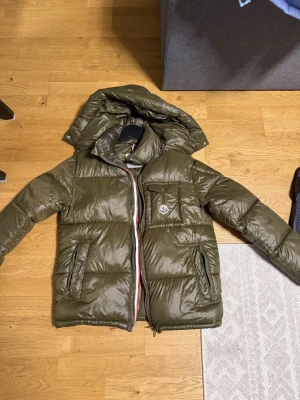  pufferjacka från Moncler - Säljer en olivgrön pufferjacka från Moncler med huva och klassisk logga på bröstet. Jackan har dragkedja framtill, fickor med dragkedja och en quiltad design som ger en schysst look. Perfekt för kalla dagar och riktigt snygg till streetwear.