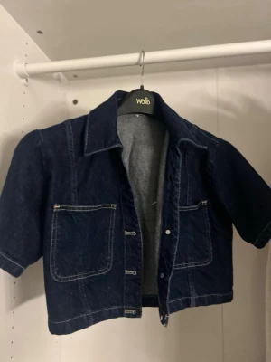 Mörkblå croppad jeansjacka - Säljer en mörkblå croppad jeansjacka från Wallis med korta ärmar och två stora fickor framtill. Jackan har tydliga vita sömmar och klassisk krage. Perfekt för dig som vill ha en chill och trendig look.
