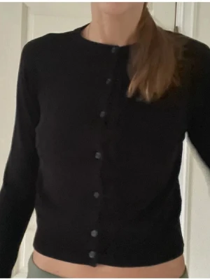 Svart kofta från H&M, storlek S - En enkel och stilren svart kofta från H&M i storlek S. Koftan har rund halsringning, långa ärmar och knappar hela vägen framtill. Perfekt att slänga över en topp eller bära stängd för en clean look.