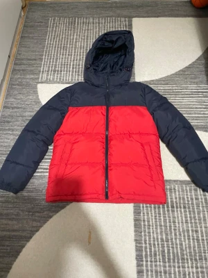 Röd och marinblå pufferjacka från Alpes - Snygg pufferjacka från Alpes i röd och marinblå färg med huva och hel dragkedja. Jackan har två sidofickor, elastiska muddar och ett coolt Alpes-märke på ärmen. Materialet är polyester och jackan är perfekt för kalla dagar. Bra vinter jacka för barn! Storlek 146. Har andvänts ca 1 gång.