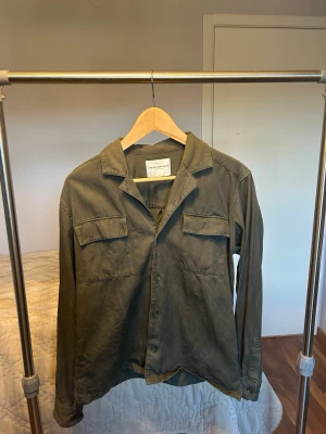 Olivgrön overshirt från These Glory Days - Olivgrön overshirt med klassisk krage och två stora bröstfickor med lock. Skjortan har knappar framtill och långa ärmar med knapp vid manschetten. Materialet är mjuk bomull och passformen är normal. Perfekt att slänga på över en t-shirt.