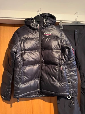 Svart Peak Performance dunjacka - Svart dunjacka från Peak Performance med huva och dragkedja framtill. Jackan har två sidofickor med dragkedja och en extra ficka på ärmen med rosa detaljer. Puffad design och logga på bröstet. Perfekt för kalla dagar och snygg till streetwear.