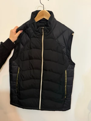 Svart dunväst från Hugo Boss - Snygg svart dunväst från Hugo Boss med quiltad design och guldfärgade detaljer vid fickorna och på ryggen. Västen har hög krage, dragkedja framtill och diskret logga på ryggen. Perfekt för lager-på-lager och streetstyle.