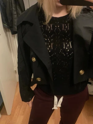 Svart croppad kavaj med guldknappar - Säljer en svart croppad kavaj med bred krage och snygga guldknappar framtill. Jackan har en loose passform och är tillverkad i ett lätt syntetmaterial. Perfekt för dig som vill ha en cool och trendig look med lite edge.