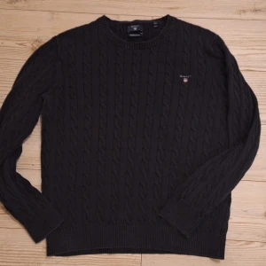 Svart kabelstickad tröja från GANT - Svart kabelstickad tröja från GANT med rund hals och diskret logga broderad på bröstet. Tröjan har långa ärmar och ribbade muddar vid ärmslut och nederkant. Perfekt för dig som gillar klassisk och stilren design. Står 2XL fast är som L