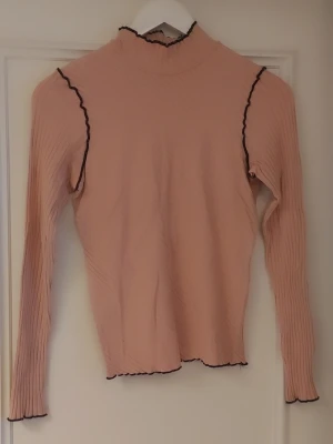 Ribbad rosa polotopp från Zara - Säljer en ribbad ljusrosa polotopp från Zara med långa ärmar. Toppen har svarta vågiga kanter vid hals, ärmslut och nederkant som ger en snygg kontrast. Materialet är stretchigt och lätt, perfekt för lager på lager.