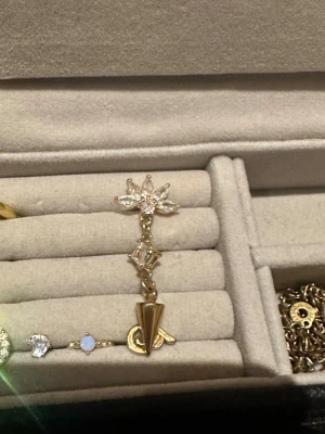 Guldglittrig örhänge med kristaller - Säljer ett snyggt örhänge i guld med glittrande kristaller formade som en blomma upptill, en fyrkantig kristall i mitten och en liten konformad detalj längst ner. Perfekt för dig som gillar unika smycken med lite edge. Endast ett örhänge! 