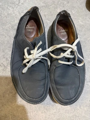 Blå moka skor från Clarks 1825 - Blå skinnskor från Clarks 1825 med vit snörning och kontrasterande vita sömmar. Skorna har rund tå och platt sula, vilket gör dem riktigt chill och klassiska. Perfekt för dig som gillar enkel och stilren design.