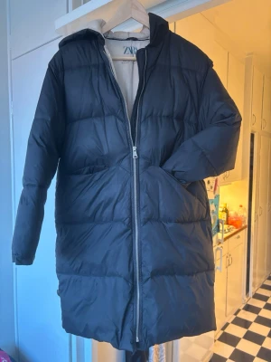 Svart lång pufferjacka från Zara - Säljer en svart pufferjacka från Zara med huva och lång passform. Jackan har en kraftig dragkedja framtill och är fodrad med ljus insida. Perfekt för kalla dagar och har en clean, modern look.