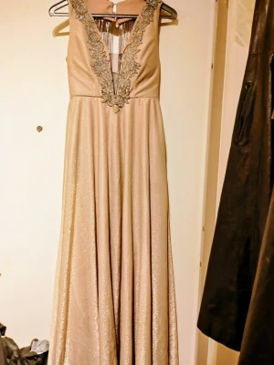 Champagnefärgad långklänning med pärlor - Säljer en elegant champagnefärgad långklänning med glittrande tyg och djup v-ringning. Klänningen har vackra broderade detaljer med pärlor runt halsringningen och långa pärlfransar i ryggen. Perfekt för dig som vill sticka ut med en lyxig look. Bal/Fest/Brudklänning köpte den för 2400:-