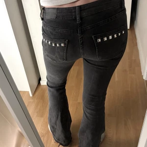 Svarta bootcut jeans med nitar - Säljer ett par svarta jeans med bootcut-snitt och coola silvriga nitar på fickorna både fram och bak. Jeansen har låg midja och är gjorda i ett klassiskt jeansmaterial. Perfekta för dig som gillar detaljer och vill sticka ut lite extra. Lagt nitarna själv