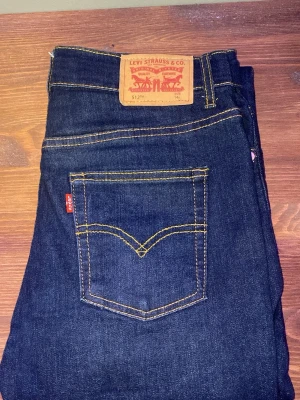 Levi's 512 mörkblå jeans straight - Mörkblå Levi's 512 jeans med klassisk gul kontrastsöm och femficksmodell. Raka ben och normal passform, tillverkade i slitstarkt denim. Märkespatch bak i midjan och metallnitar vid fickorna för extra detaljer. Storlek: 32