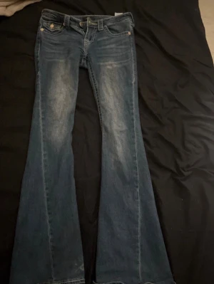 True Religion bootcut jeans  - Säljer ett par blå jeans från True Religion med bootcut-snitt och klassisk gul kontrastsöm. Jeansen har slitningar och detaljerade bakfickor med knappstängning och broderade sömmar. Materialet är jeans, dom är lite slitna nere vid fötterna (storlek 24)