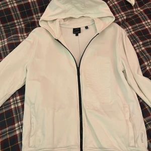 Armani Exchange Hoodie - Vit hoodie från Armani Exchange. Från utsidan har den knappt några synliga tecken på användning. Däremot är den tunn och ganska hård i insidan.