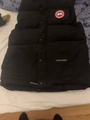 Svart dunväst från Canada Goose - Svart dunväst från Canada Goose med klassisk logga på bröstet och tydlig märkning på fickan. Västen har en quiltad design, knappar och dragkedja framtill. Perfekt för kalla dagar och har en stilren look som funkar till många outfits. Passar storlek S bra! Samt M. ÄKTA VÄST