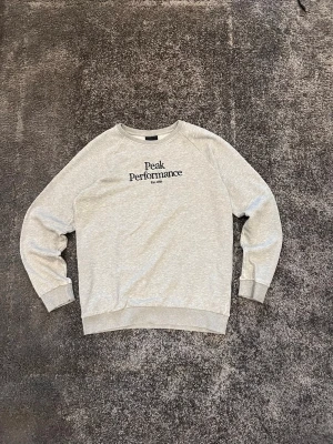 Grå Peak Performance sweatshirt - Säljer en grå sweatshirt från Peak Performance med svart broderad logga på bröstet. Tröjan har rund hals, ribbade muddar vid ärmslut och nederkant samt långärmad passform. Perfekt för chill dagar och enkel att matcha med jeans eller joggers. Tröjan har en slitning i på slutet av armarna som visas på bilderna. Det är nog lätt att fixa om man är lite händigare än mig! Skriv om du undrar något!☺️