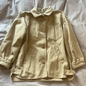 Beige vårjacka från Zara - Säljer en beige jacka från Zara med klassisk krage, stora knappar och fickor fram. Perfekt för våren, hösten men även som en sommarjacka. Har ett par små fläckar.   Barnstorlek 140, men jackan passar även dam stl. XS! Ärmarna blir då något kortare.
