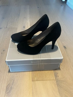 Svarta pumps från Tamaris, storlek 39 - Snygga svarta pumps från Tamaris i storlek 39. Skorna har rund tå, hög klack och är tillverkade i mocka med en mjuk insida. Perfekta för dig som gillar klassisk stil med en modern touch. Säljes med originalkartong.