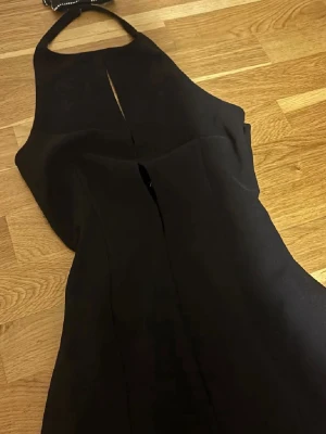 Svart kort klänning med rosett - Säljer en svart kort klänning med halterneck. Klänningen har en dekorativ rosett med pärldetaljer i halsen och en tight passform som framhäver figuren. Perfekt för dig som vill sticka ut med en elegant och stilren look.