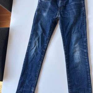 Levi's  Jeans Blå - Säljer ett par Levi's jeans i klassisk blå tvätt med snygga slitningar och kontrastsömmar. Modellen har fem fickor, knapp och dragkedja fram samt Levi's patch och röd etikett bak. Perfekt passform med smal siluett och stretchigt jeanstyg. Tyvär ett litet hål framme vid gylfen men lättlagat och man ser knappast att det finns när man använder dem