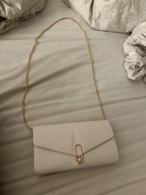 Beige kuvertväska med kedja - Snygg beige kuvertväska med flätat mönster och guldfärgad kedja som axelrem. Väskan har ett elegant spänne i guld på framsidan och är perfekt för att lyfta din outfit. Materialet känns som skinn och designen är stilren och modern.Den är även helt ny med prislappen kvar ifrån anna field