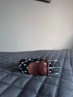 Svart vitt grått flätat bälte - Tja det är helt nytt från Bältgallerian. Det är 105cm långt och 3,5cm brett. Det är i ett elastisk material som passar större storlekar.