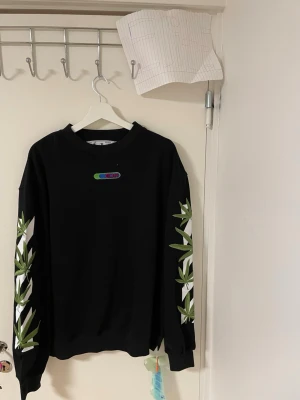 Svart Off-White sweatshirt  - Svart sweatshirt från Off-White i storlek L med gröna bladprint längs ärmarna och stor vit grafisk logga på ryggen. Rund hals och ribbade muddar. Materialet är mjuk bomull och passformen är normal. Perfekt för dig som gillar streetwear och vill sticka ut.Liknande modell som orginala. 