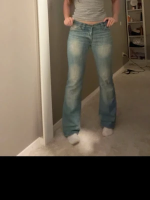 Bootcut ljusblå jeans med slitningar - Säljer ett par ljusblå bootcut jeans med snygga slitningar och tvättade detaljer. Jeansen har klassisk femficksdesign, låg midja och är tillverkade i jeansmaterial med en skön passform. Perfekt för dig som gillar retrovibbar och vill ha ett par jeans som sticker ut.