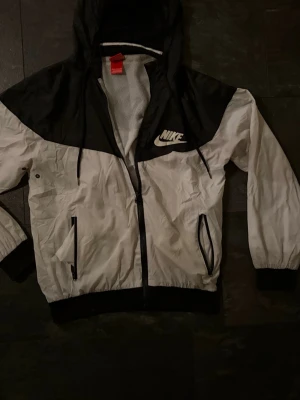 Svartvit vindjacka från Nike - Nike vindjacka i svart och vitt med huva och meshfoder. storlek L men den är M i storlek. Jackan har dragkedja framtill, två fickor med dragkedja och justerbar snörning i huvan. Klassisk logga på bröstet och elastiska muddar vid ärmar och midja. Perfekt för dig som gillar sportig stil. Inga defekter har märkts. Kontakta vid frågor och funderingar!
