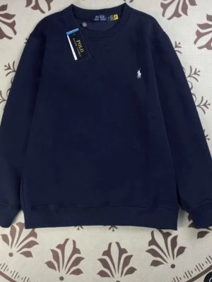 Marinblå sweatshirt från Polo Ralph Lauren - Klassisk marinblå sweatshirt från Polo Ralph Lauren med den ikoniska vita logon broderad på bröstet. Tröjan har rund halsringning och långa ärmar, perfekt för en clean och stilren look.
