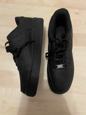 Nike Air Force 1 helsvarta sneakers - Säljer ett par klassiska Nike Air Force 1 i helsvart färg. Skorna har en låg siluett, rund tå och snörning framtill. Tillverkade i skinn med perforerade detaljer på tån för extra ventilation. Snygg metallbricka med 'AF1' på snörena och ikonisk sula.