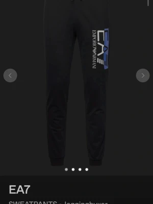 Svarta mjukisbyxor från EA7 Armani - Snygga svarta mjukisbyxor från EA7 Emporio Armani med stort tryck i vitt och blått längs ena benet. Byxorna har dragsko i midjan och ribbade muddar vid bensluten. Perfekta för chill eller träning. Nypris runt 1000kr