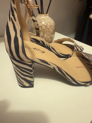 Zebramönstrade sandaler med klack - Snygga sandaler från Alley i storlek 37 med zebramönster i svart och beige. Skorna har en bred rem över tårna, öppen tå och en stadig blockklack. Justerbart spänne runt vristen och mockaliknande material ger en trendig look.