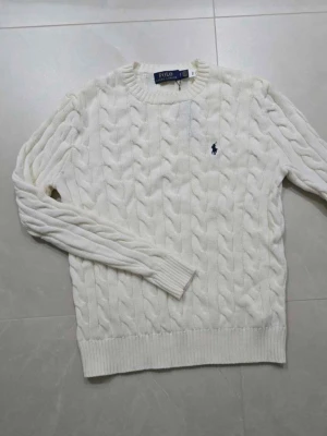 Vit kabelstickad tröja Polo Ralph Lauren - Kabelstickad vit tröja från Polo Ralph Lauren med klassisk blå logga på bröstet. Rund halsringning och ribbade muddar vid ärmslut och nederkant. Perfekt för dig som gillar en clean och tidlös look.