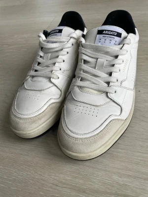 Vita sneakers från Axel Arigato strl 42 i mycket bra skick  - Säljer ett par stilrena vita sneakers från Axel Arigato i storlek 42. Skorna har en låg profil, rund tå och platt sula. Ovandelen är i skinn med detaljer i mocka och perforeringar på sidan och tån. Svart häl och vita snören ger en clean look. Skicket är mycket fint. Typ oanvända. 