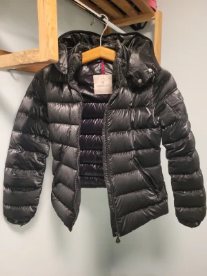 Svart Moncler pufferjacka med huva - Säljer en svart pufferjacka från Moncler med glansig finish och huva. Jackan har dragkedja framtill, två sidofickor med dragkedja och klassisk Moncler-logga på insidan. Perfekt för kyliga dagar och riktigt snygg streetstil. Storlek 12A/xs-s. Den passar som xs
