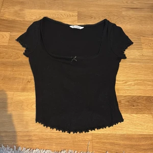 Svart topp med rosett  - Säljer en svart topp från Miss other stories med korta ärmar och söt liten rosett framtill. Toppen har vågiga kanter vid ärmslut och nederkant samt en fyrkantig ringning med spetsdetalj. Materialet är mjukt och stretchigt, perfekt till många olika looks.