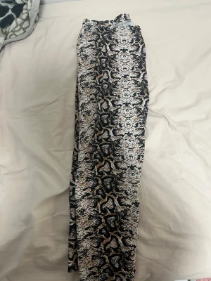 Ormmönstrade byxor med resårmidja - Säljer ett par snygga byxor med ormmönster i svart, beige och vitt. Byxorna har resår i midjan och är raka i modellen, perfekta för dig som vill sticka ut lite extra. Materialet känns lätt och luftigt, passar bra till olika outfits.