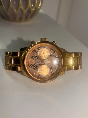 Guldig klocka från Guess - Säljer en snygg guldig klocka från Guess med metallarmband och flera urtavlor. Klockan har detaljer i guld och romerska siffror på urtavlan. Perfekt accessoar för att lyfta din stil.
