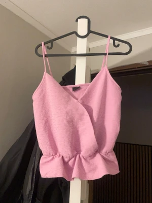 Rosa omlottopp från Gina Tricot - Säljer en ljusrosa omlottopp från Gina Tricot i storlek S. Toppen har smala axelband, volangkant nedtill och en snygg omlottdesign framtill. Perfekt för varma dagar och enkel att styla med jeans eller kjol.