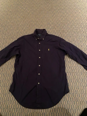 Mörkblå skjorta från Ralph Lauren - Klassisk mörkblå skjorta från Ralph Lauren i bomull med button-down krage och broderad gul logga på bröstet. Skjortan har lång ärm, normal passform och vita knappar framtill. Perfekt till jeans eller chinos för en clean look.