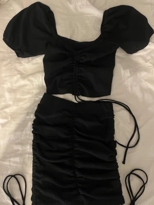 Ett svart sett från Gina Tricot  - Säljer ett svart sett från Gina Tricot. En topp med snörning framtill. Toppen har en tight passform och är sydd i ett mjukt, lätt stretchigt material men som sitter perfekt på kroppen. Kjolen till har snörning på sidorna och scrunch fram & baktill för att skapa en snyggare passform. Använt någon enstaka gång. 