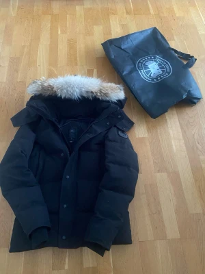  Canada Goose - Säljer en svart dunjacka från Canada Goose med fluffig päls på huvan. Jackan har dragkedja och knappar framtill, samt ribbade muddar vid ärmsluten. Perfekt för kalla vinterdagar och riktigt snygg streetstil. Allt og ingår tags osv 