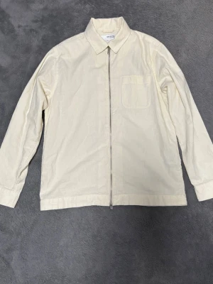 Beige overshirt från Selected Homme - Snygg beige overshirt från Selected Homme med rak passform och klassisk krage. Jackan har en bröstficka, långa ärmar med knapp vid ärmslut och dragkedja framtill. Perfekt att slänga över en t-shirt för en clean och enkel look.
