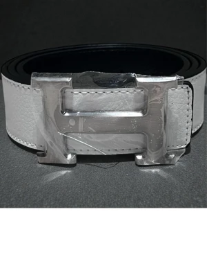 Vit skinnväst med H-spänne - Säljer en stilren vit väst med ett stort H-format metallspänne framtill. Västen är gjord i slätt skinn med diskreta sömmar längs kanterna och har en klassisk, rak form. Perfekt för dig som gillar minimalistisk design och vill ha en cool detalj i din outfit.